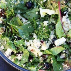 A corner of a glass bowl of quinoa salad with feta, fennel and blueberries from above on a black background