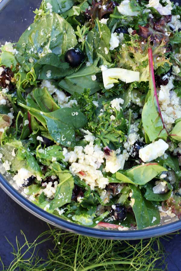 A corner of a glass bowl of quinoa salad with feta, fennel and blueberries from above on a black background