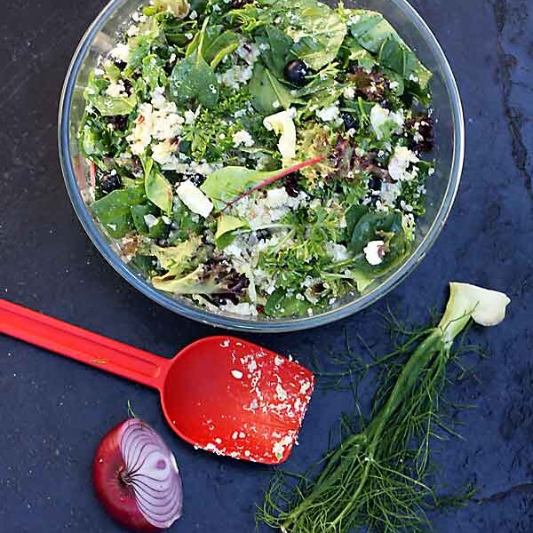 A glass bowl of quinoa salad with feta, fennel and blueberries on a black background from above and with a red plastic spoon and veggie scraps