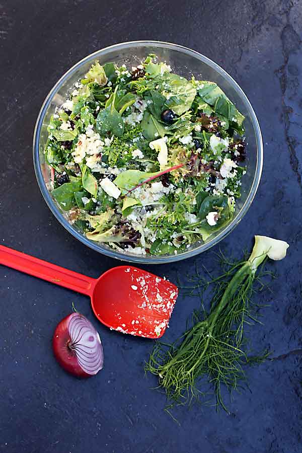 A glass bowl of quinoa salad with feta, fennel and blueberries on a black background from above and with a red plastic spoon and veggie scraps