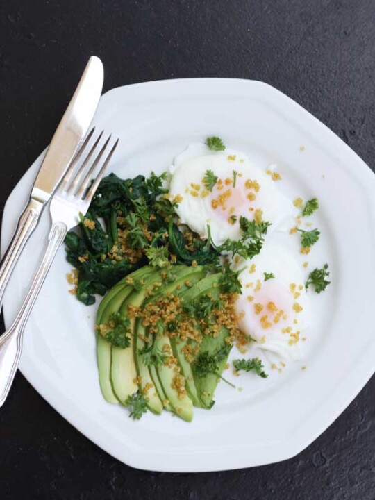 Poached eggs with avocado, spinach & crispy quinoa on a white plate with a knife and fork and on a black background