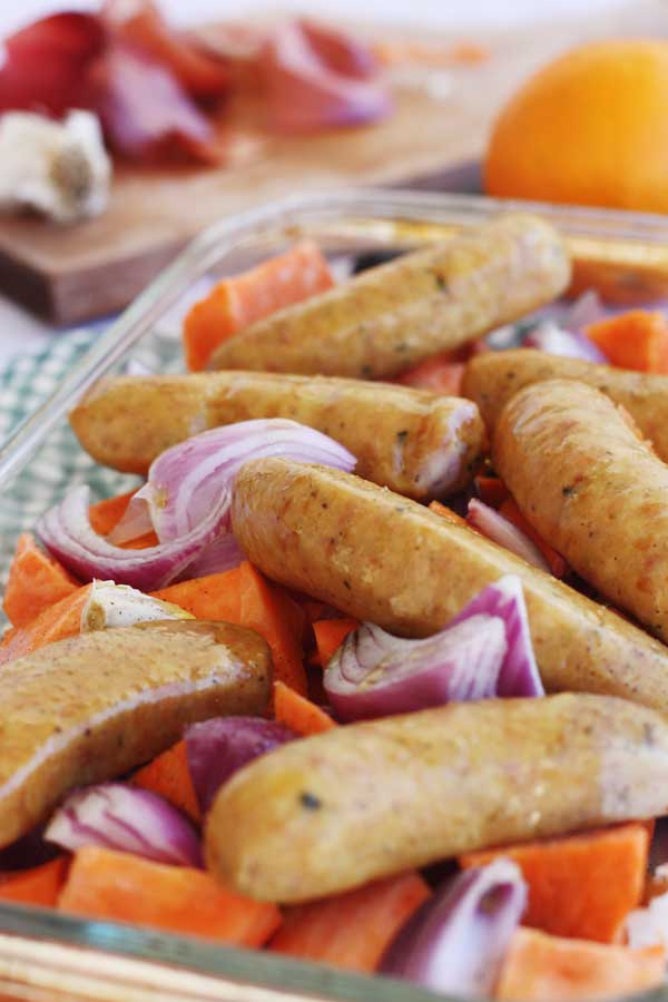 closeup of sausage, sweet potato and orange tray-bake on a green checked background ready to go in the oven.