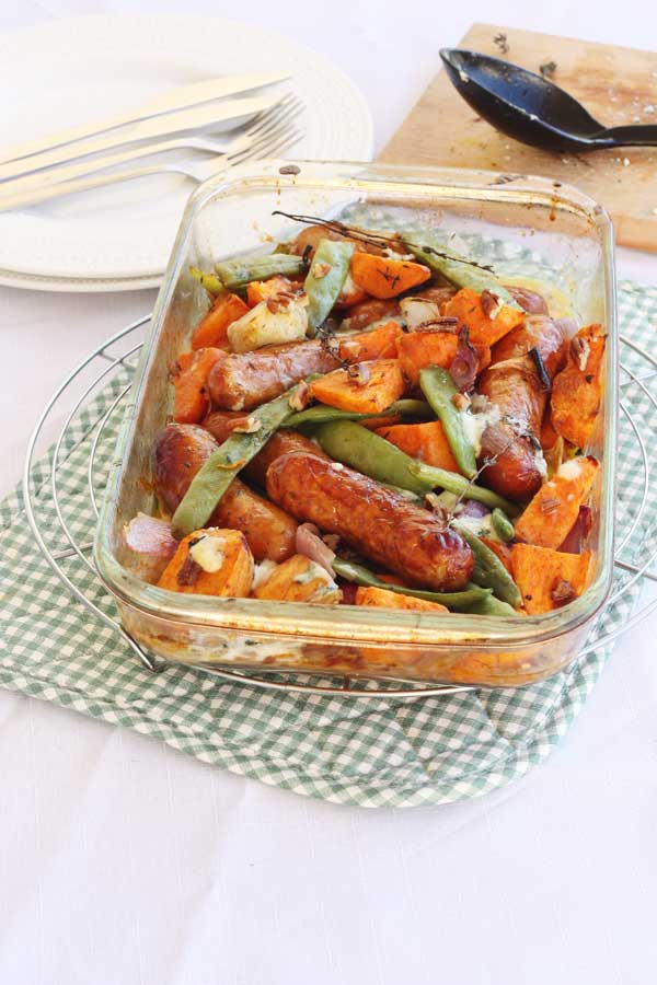 Sausage, sweet potato and orange tray-bake in a glass baking dish just out of the oven on a green and white checked place mat.