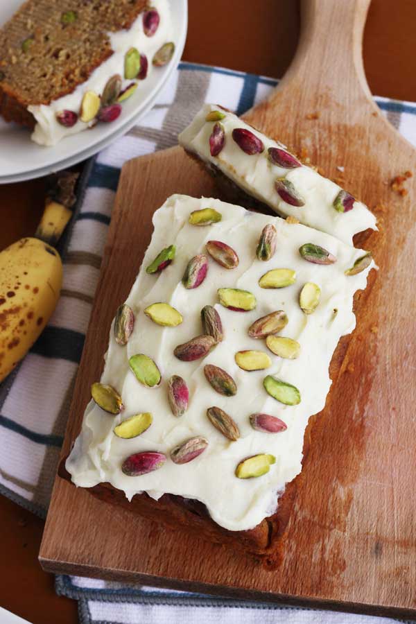 banana pistachio loaf with white chocolate cream cheese frosting from above on a wooden board