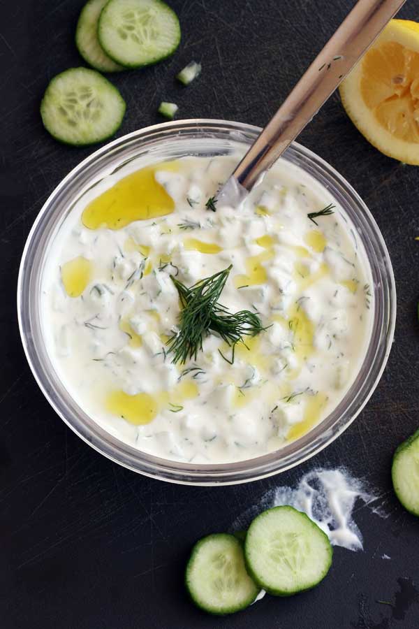 A bowl of tzatziki from above with olive oil and dill on top and with a spoon in it and lemon and cucumber scattered around.