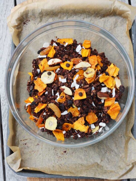 Bowl of tropical chocolate granola on a baking sheet lined with brown baking paper from above.
