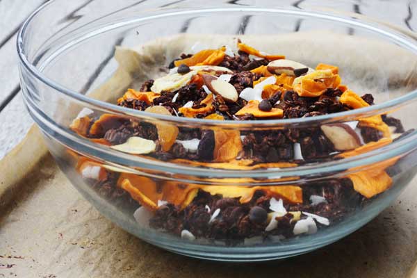 A glass bowl of crunchy tropical chocolate granola on brown baking paper.