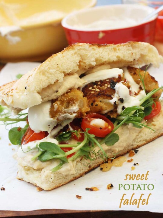 A sweet potato falafel sandwich from the side on baking paper with kitchen equipment in the background.