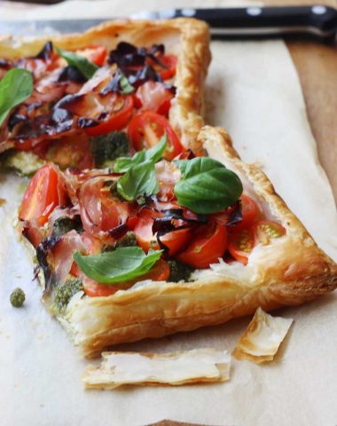 A closeup of a puff pastry tart with tomatoes on baking paper and with another piece and a knife in the background