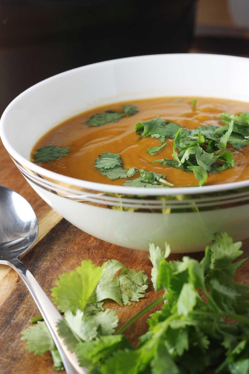 closeup of a bowl of thai pumpkin soup on a wooden board with a spoon and herbs