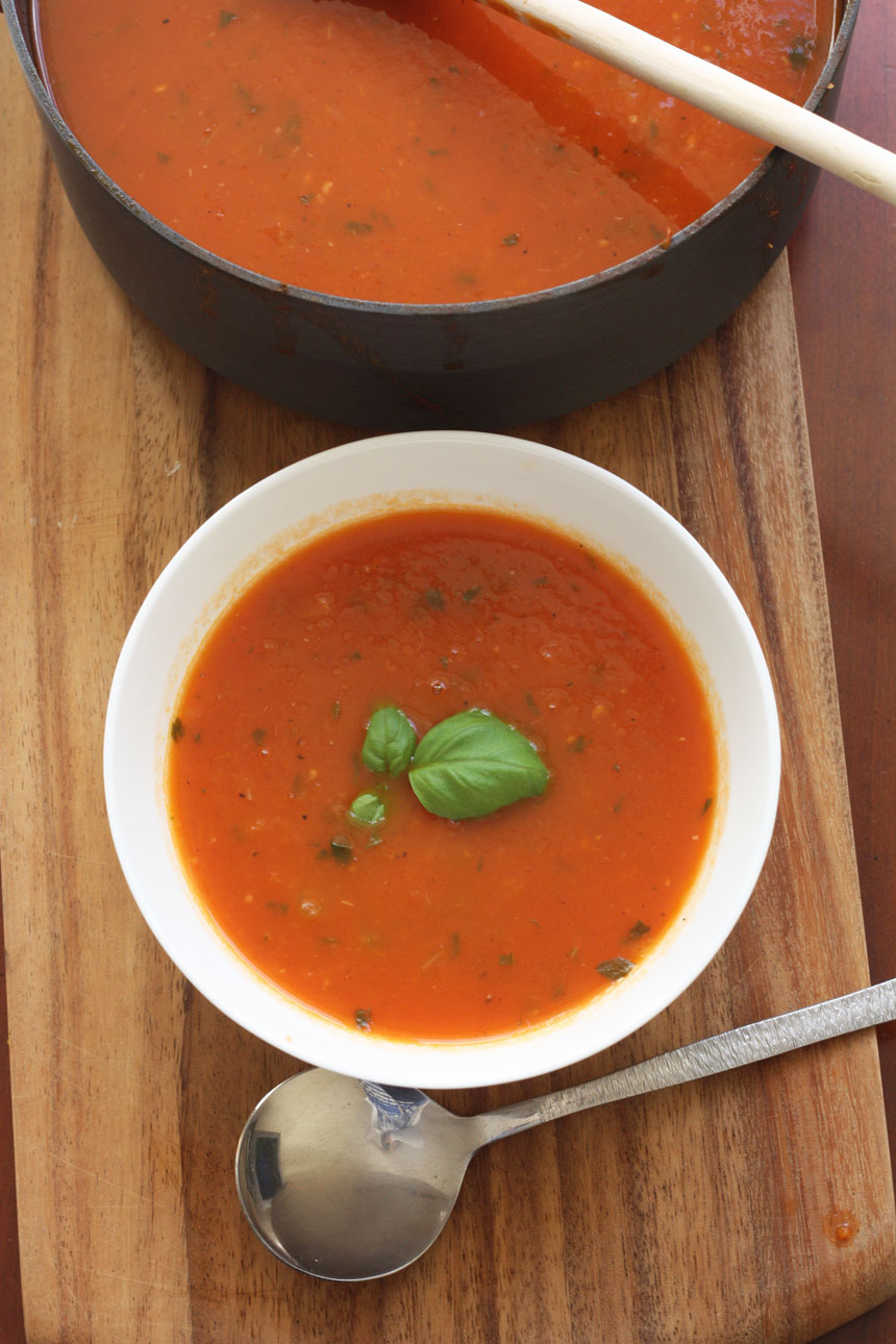A bowl of classic tomato soup on a wooden cutting board from above with a pot and a spoon next to it
