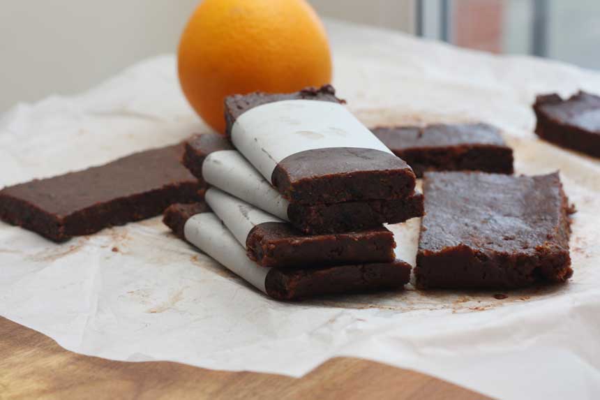 healthy chocolate orange date bars stacked on top of each other on baking paper with others around and an orange in the background.