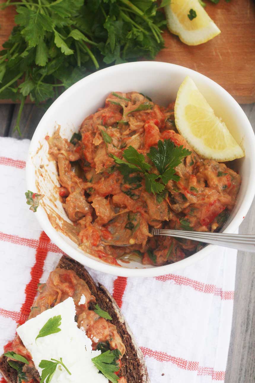 A bowl of red pepper and eggplant dip with a slice of lemon and on a red and white checked tea towel