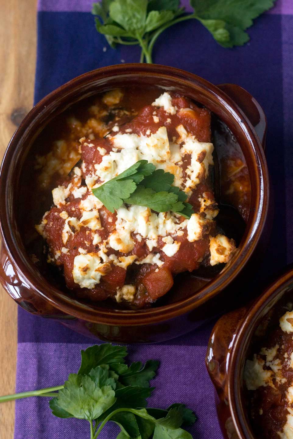 Overhead view of baked eggplant in tomato sauce in a brown ceramic pot with feta cheese on top and on a purple tea towel.