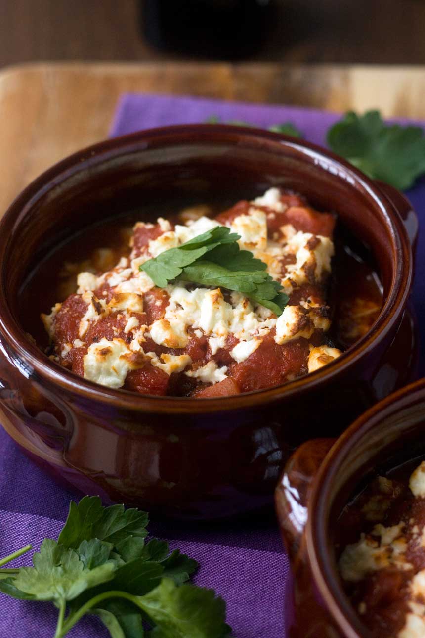 Closeup of baked eggplant covered in tomato sauce and feta cheese in a brown ceramic pot on a purple tea towel.