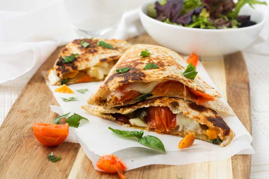 Caprese quesadillas stacked on top of each other on baking paper and a wooden board with a bowl of green salad.