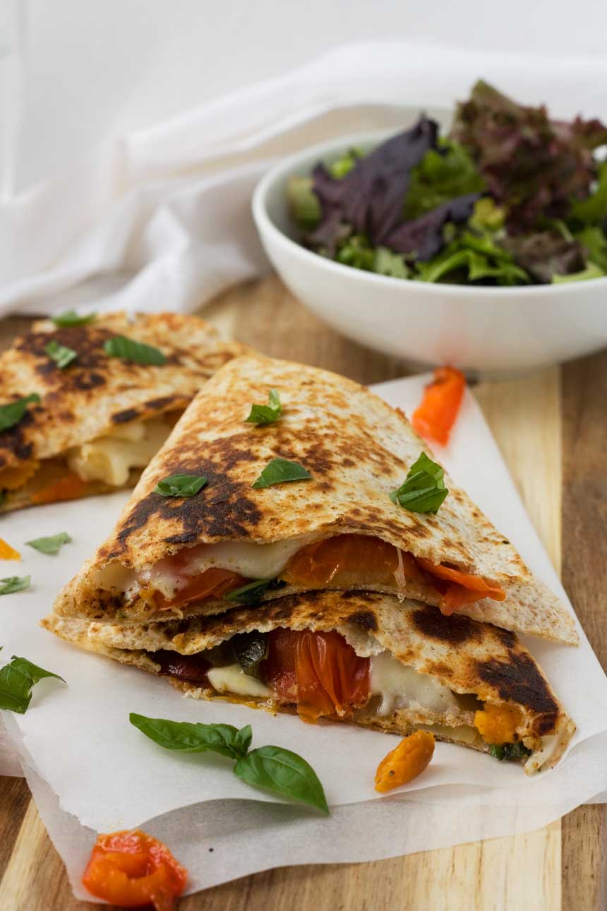 Caprese quesadillas stacked on top of each other on baking paper and a wooden board with a bowl of green salad.