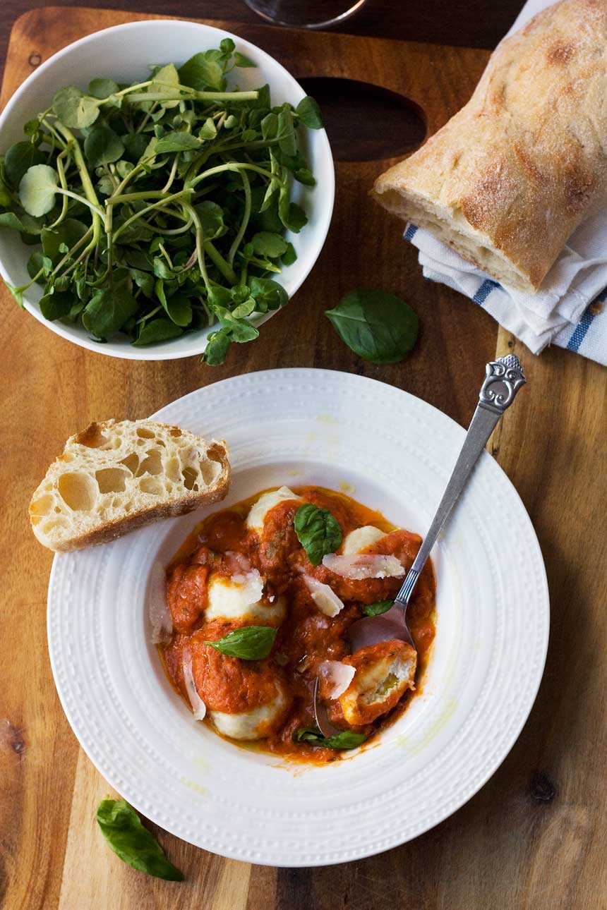 Ricotta gnudi in tomato sauce in a white bowl with a spoon with salad and bread in the background.