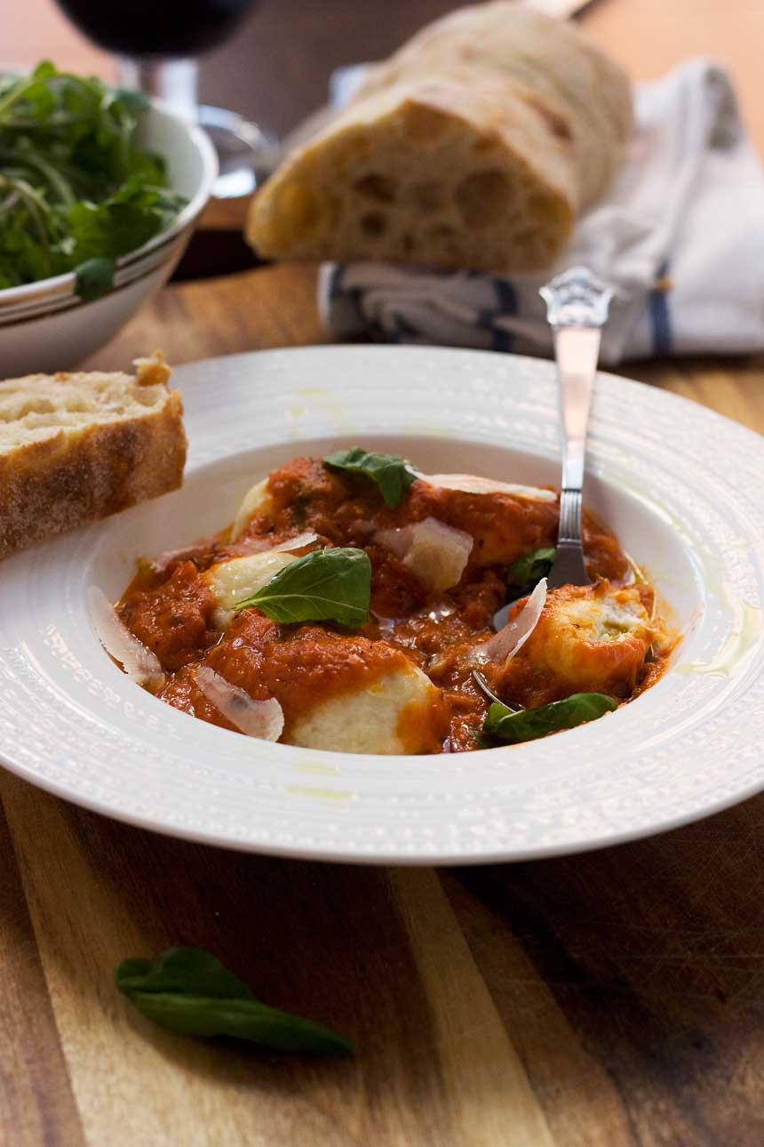 Ricotta gnudi in tomato sauce with a spoon in a white bowl with salad and bread in the background.