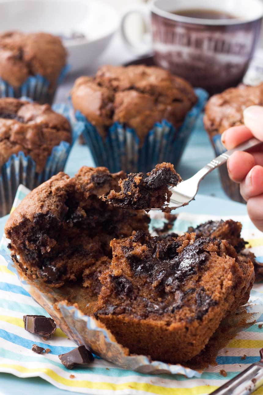 Someone eating a triple chocolate muffin with a fork - the muffin is split open so you can see the inside and there are more muffins in the background