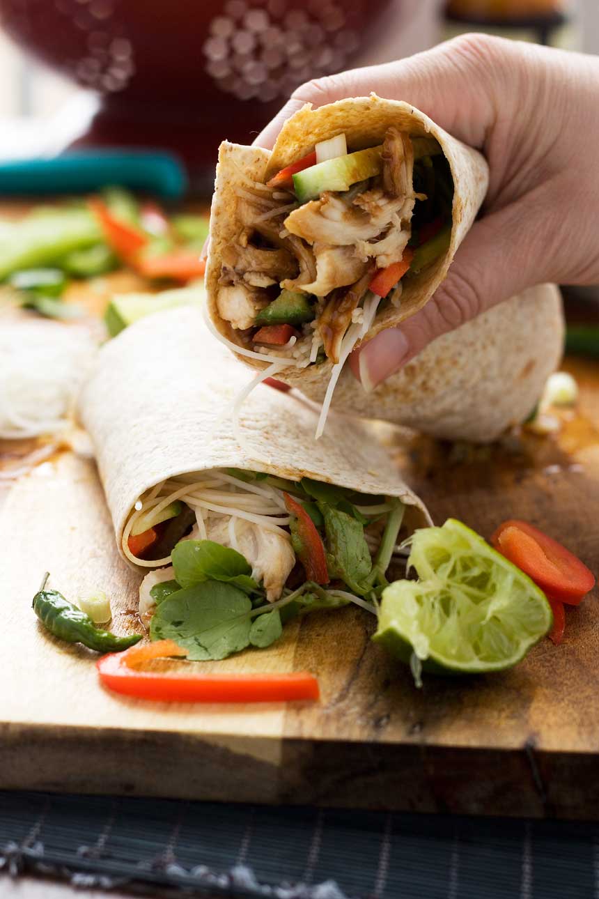 A person holding up half a hoi sin chicken noodle wrap over another half on a wooden board.