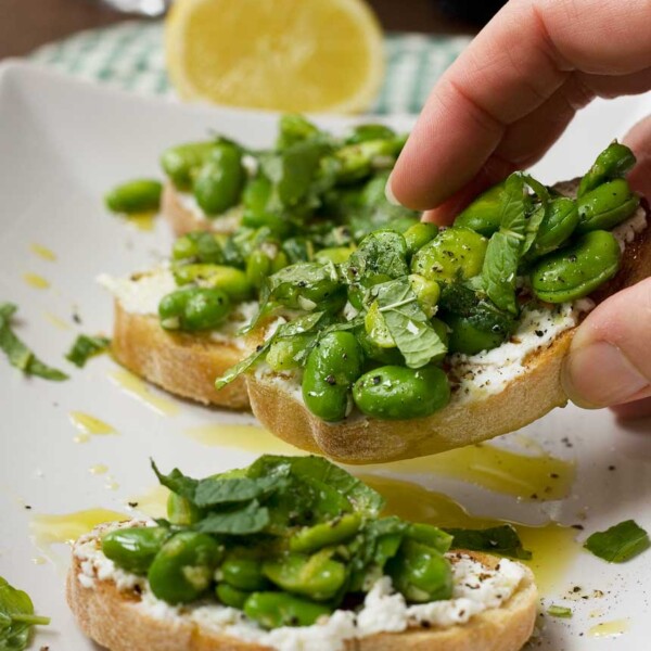 Someone taking a broad bean, mint and ricotta crostini off a plate of 4 with a lemon and checked cloth in the background