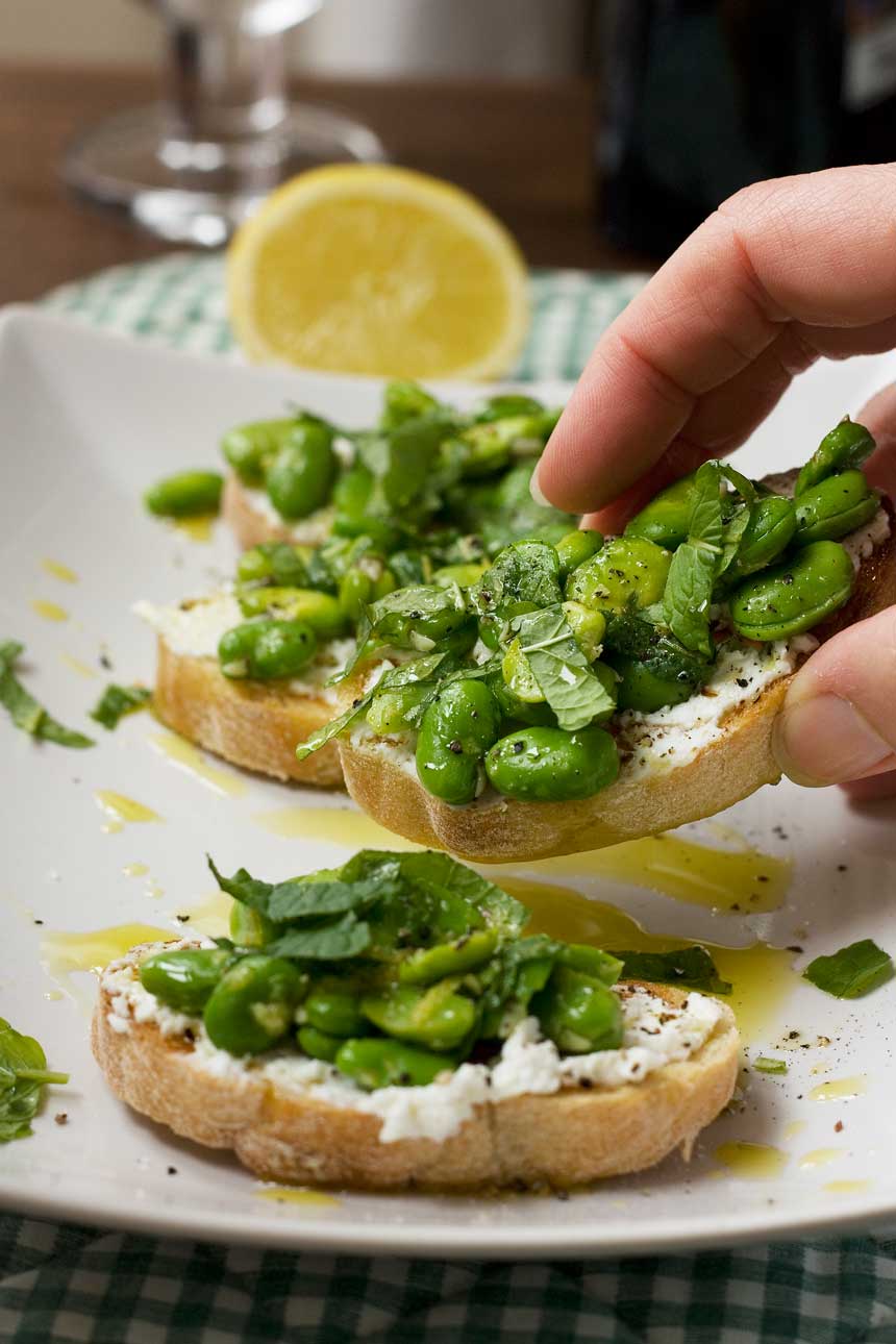 Someone taking a broad bean, mint and ricotta crostini off a plate of 4 with a lemon and checked cloth in the background