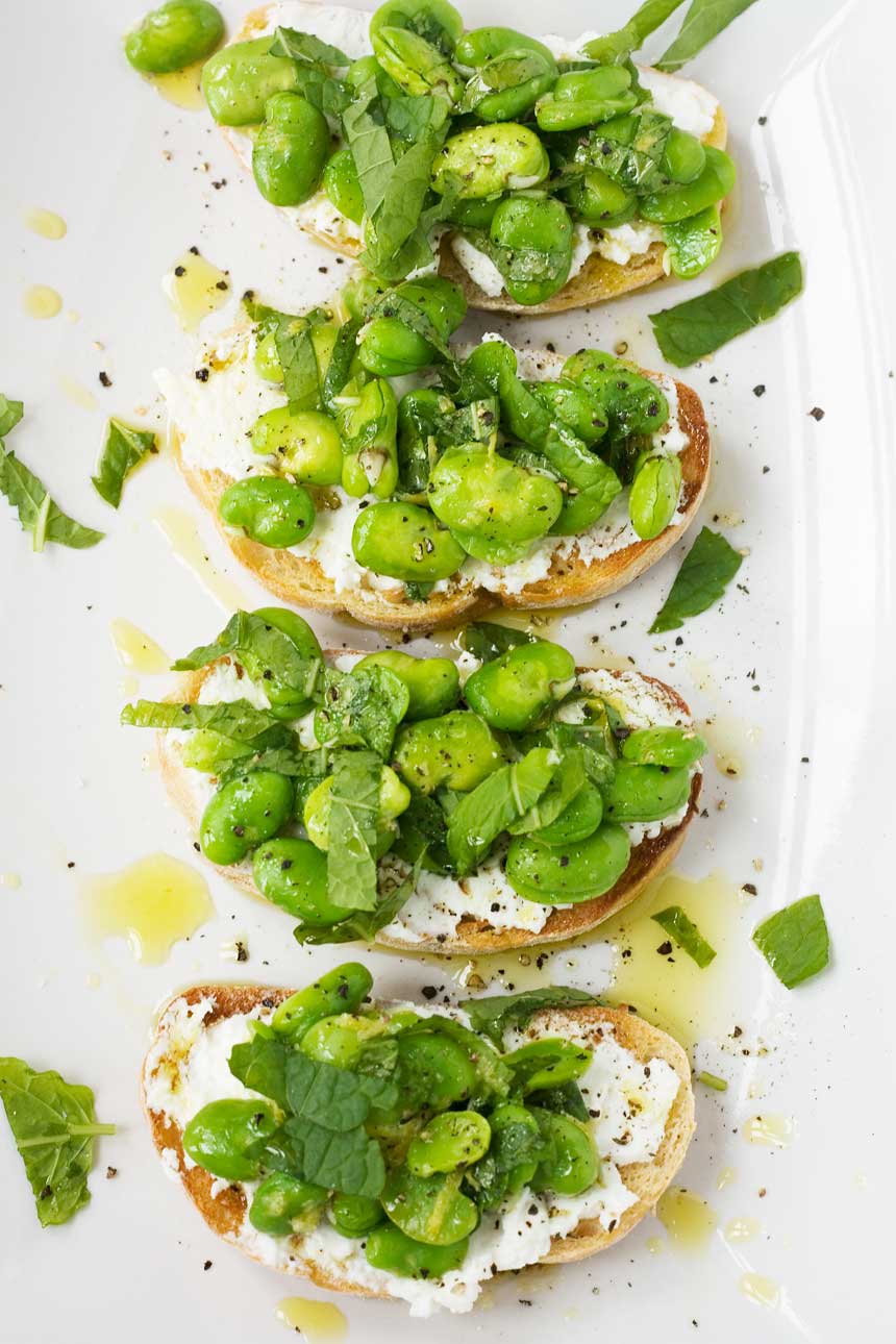 An overhead shot of 4 fava bean, mint and ricotta crostini on a white rectangular plate