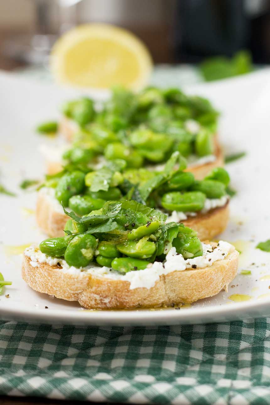 A close up of a fava bean, mint and ricotta crostini on a white rectangular plate - there are more in the background