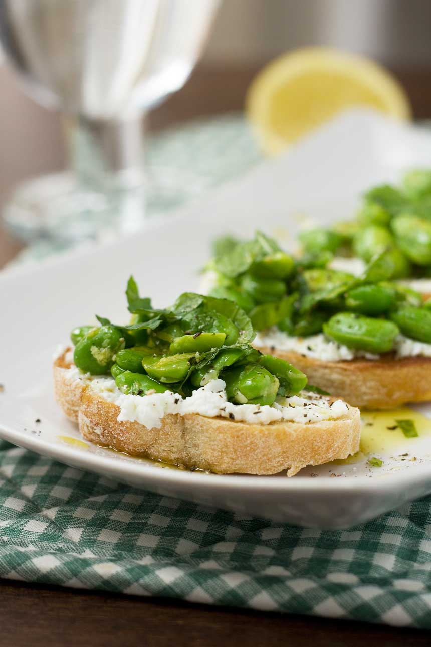 A close up of a fava bean, mint and ricotta crostini on a white rectangular plate with a green checked cloth under it
