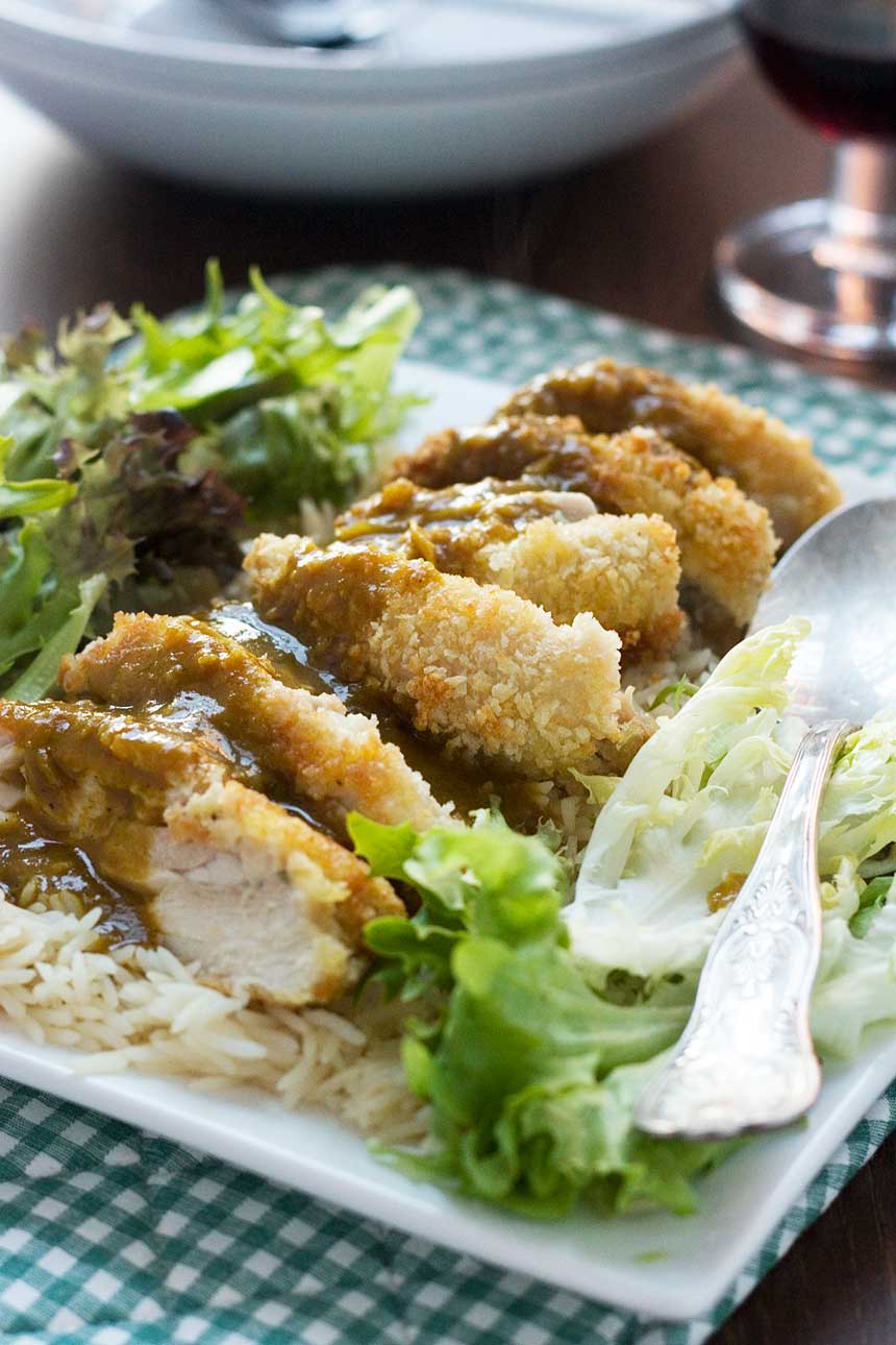 A square plate of chicken katsu and curry sauce on a bed of rice and lettuce and on a green checked cloth