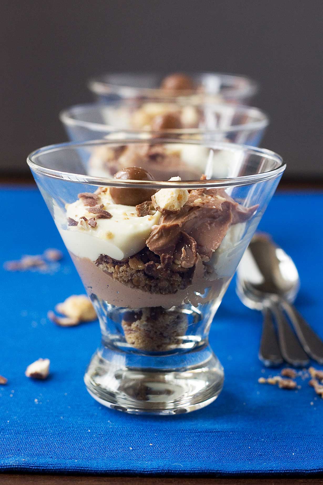 Chocolate malt cheesecakes in a glass on a blue background with spoons at the side.