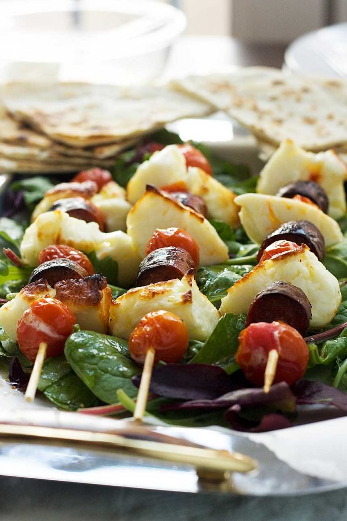 A close up of halloumi, chorizo and tomato skewers on a bed of lettuce with pitta bread in the background