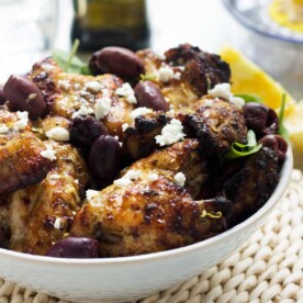 A big white dish of Greek baked chicken wings with feta and olives on top and an olive oil bottle in the background