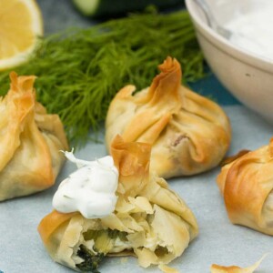 A Greek spinach and feta filo parcel with a bite out of it and a spoonful of tzatziki on it - more parcels and herbs in the background