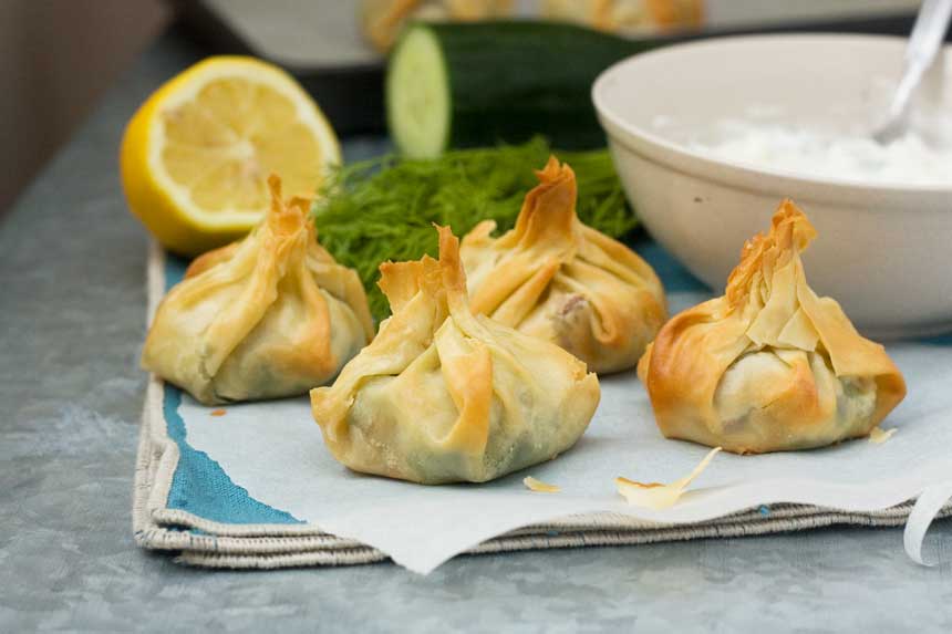 Greek filo parcels on baking paper and a blue cloth with ingredients behind.