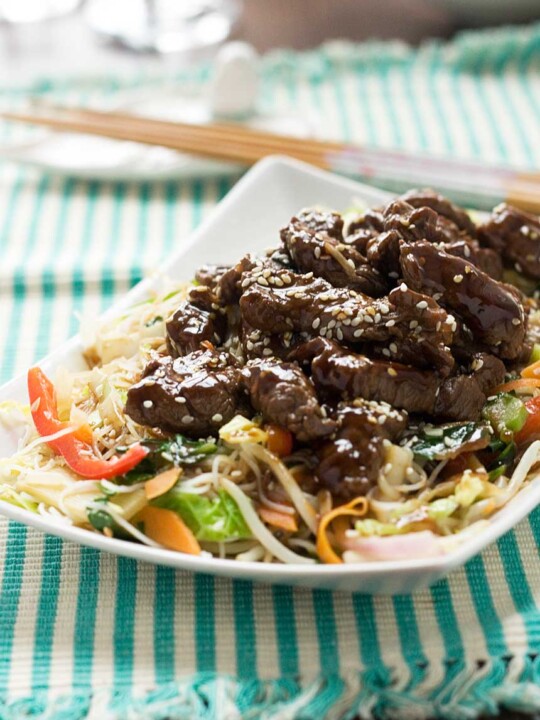 A close up of hoi sin beef and noodles on a rectangular white plate on a green and white striped cloth and with chopsticks in the background.