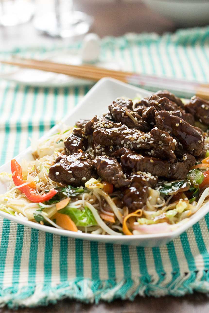 A close up of hoi sin beef and noodles on a rectangular white plate on a green and white striped cloth and with chopsticks in the background.