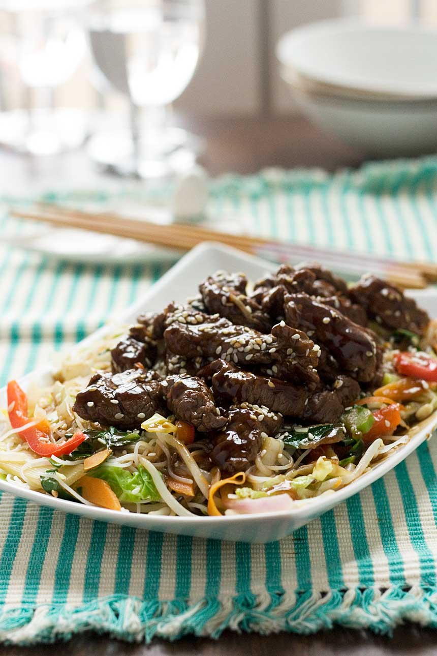 A close up of hoi sin beef and noodles on a rectangular white plate on a green and white striped cloth and with chopsticks in the background.