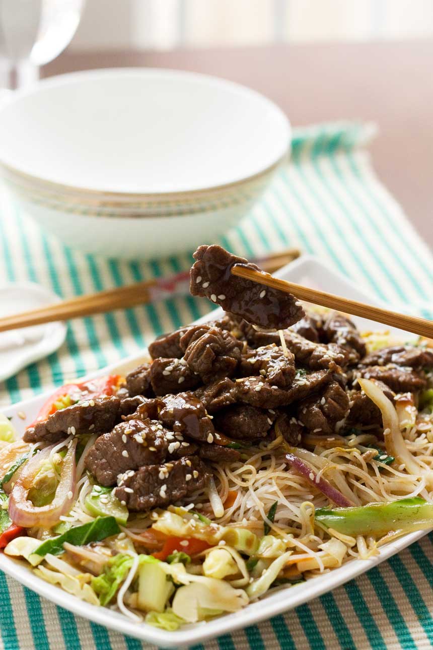 A close up of hoi sin beef and noodles on a rectangular white plate on a green and white striped cloth and with chopsticks and a bowl in the background.