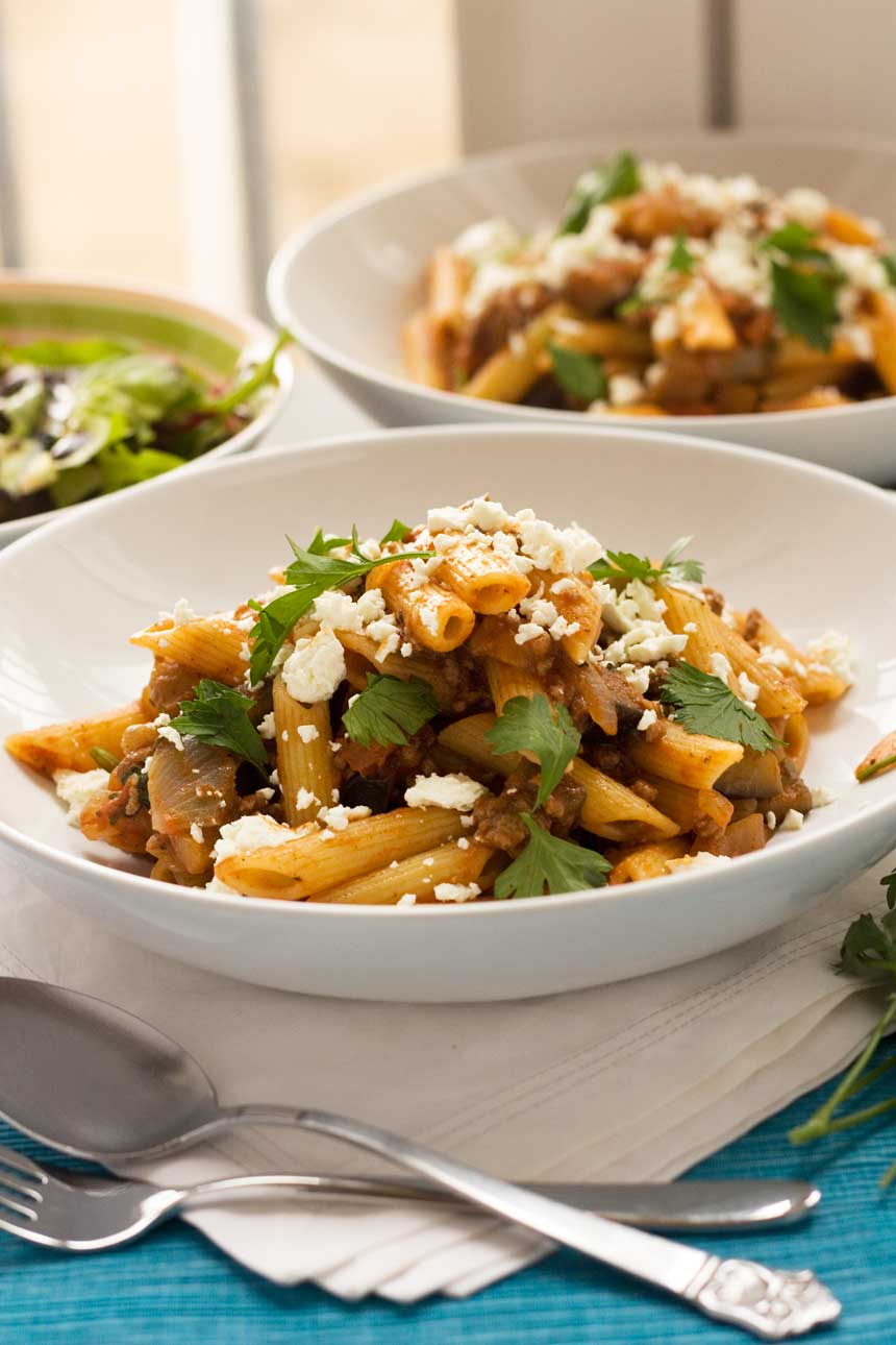 A closeup of a white bowl of penne pasta with feta on top on a white cloth and with another bowl in the background.