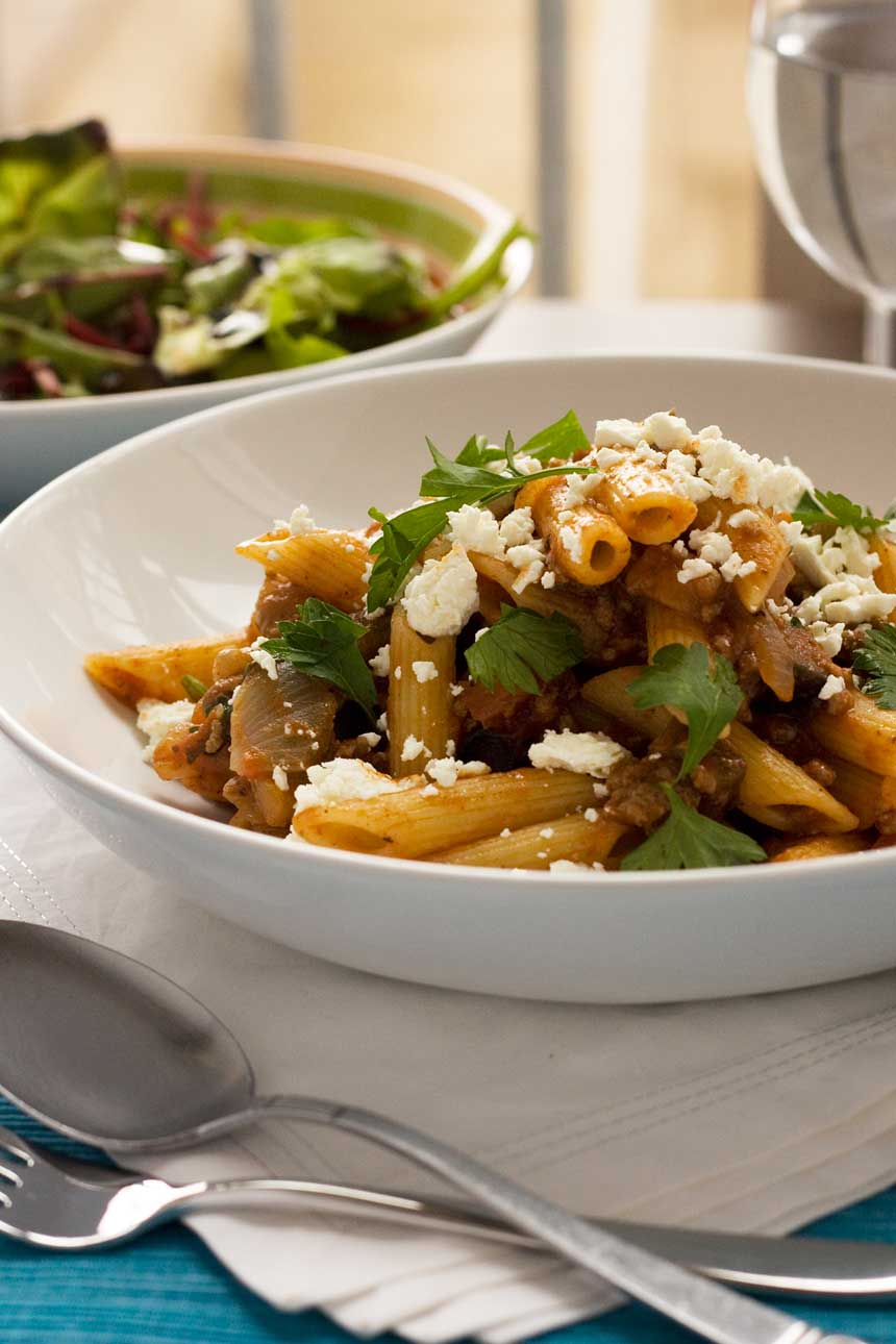 A closeup of a white bowl of penne pasta with eggplant and feta on a white cloth with another bowl in the background.