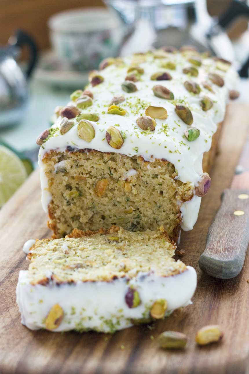 A closup of a sliced pistachio, lime and zucchini cake on a wooden board with a wooden handled knife.