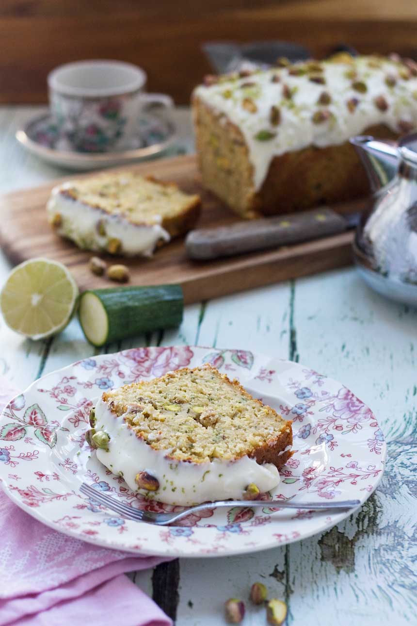 A slice of pistachio, lime and zucchini loaf on a pretty pink plate on a white wood background with more cake and ingredients in the background.