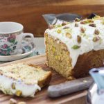 A courgette loaf with pistachios on it on a wooden board and with a pretty tea cup in the background.