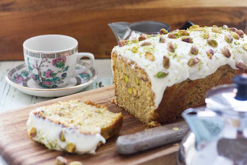 A courgette loaf with pistachios on it on a wooden board and with a pretty tea cup in the background.