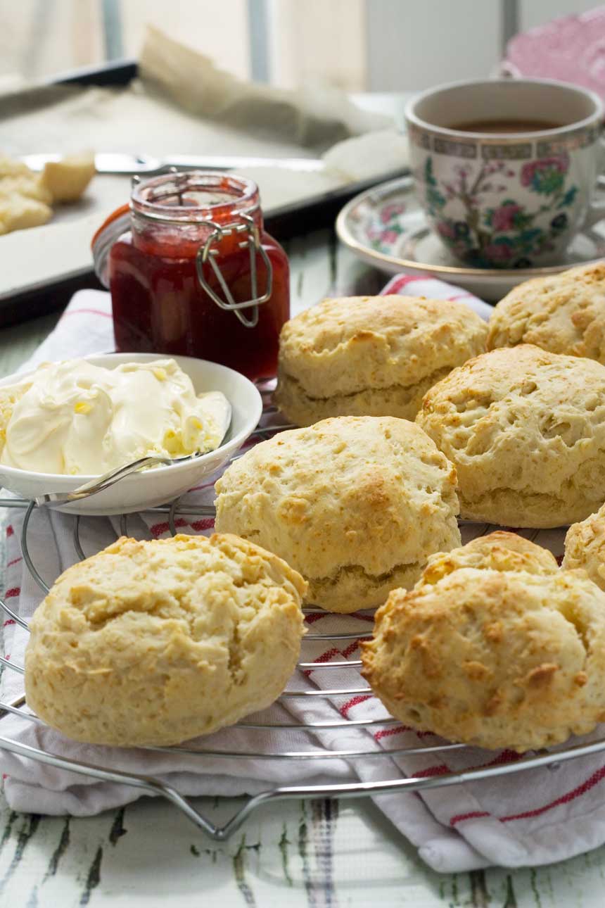 A close up of 3-ingredient scones with jam and cream on a wire rack with a pretty cup in the background.