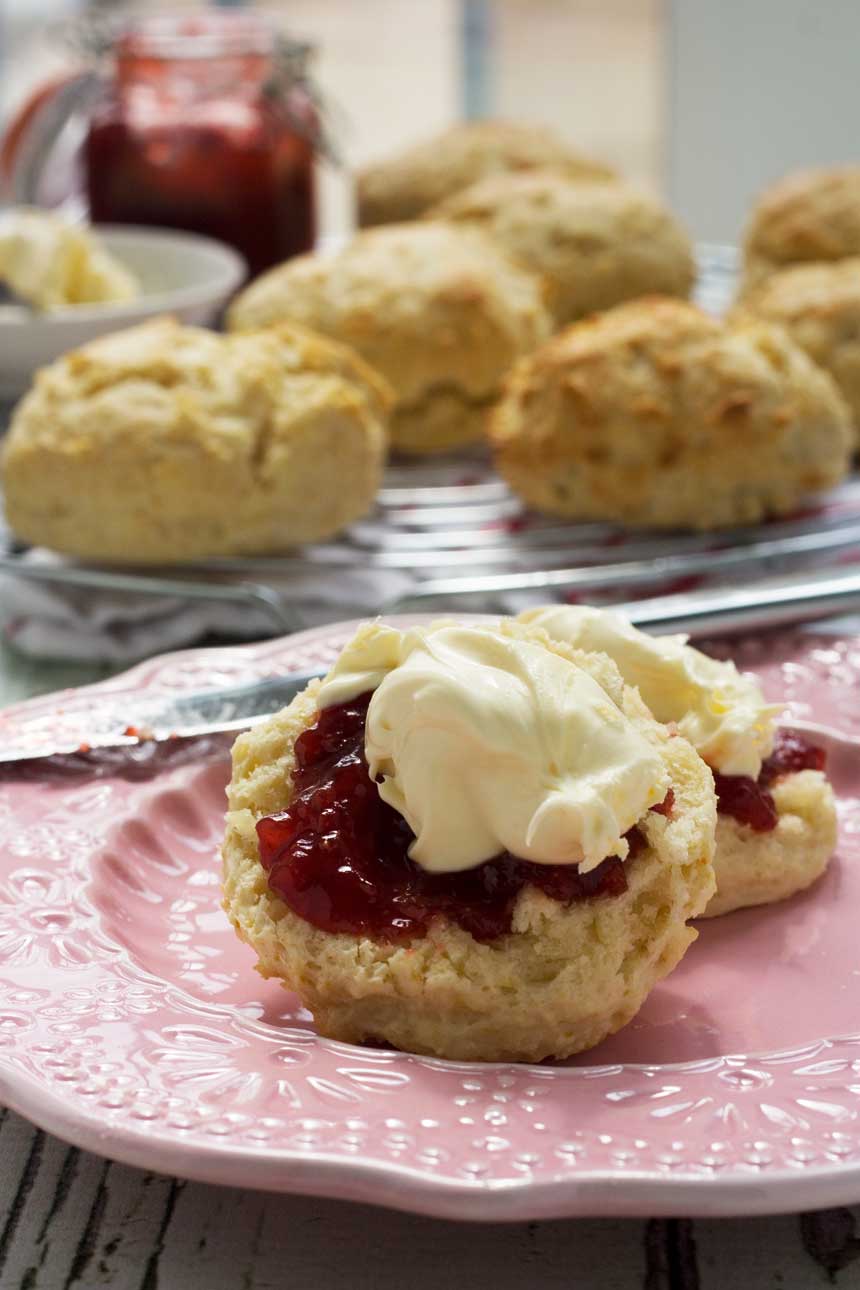 A close up of 3-ingredient scones with jam and cream on a pink plate with more scones in the background.