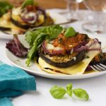 A freestyle eggplant lasagna on a white plate with a fork on a white background with a dark blue cloth and wine glasses.