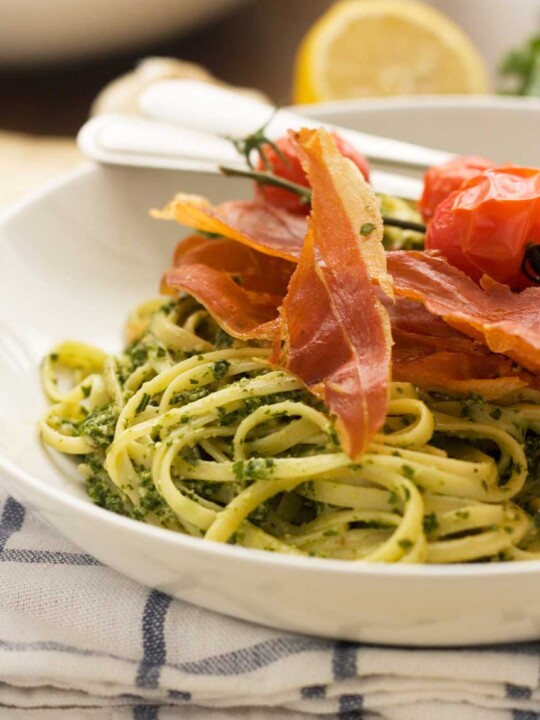 A closeup of a white bowl of kale pesto linguine with cutlery on a blue and white checked tea towel.